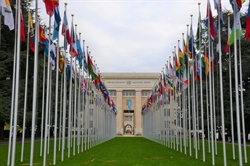 [ai] A view of the United Nations building in Geneva, framed by rows of international flags on tall poles, set against a cloudy sky.